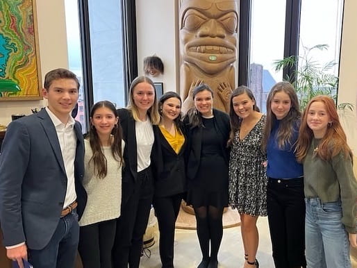 A group of eight young adults in formal attire stand in a senators office. A large wood totem with a carved face stands prominently in the background.